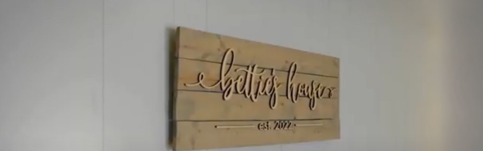 Wooden wall sign with cursive text that reads "Bettie's House," mounted on a white wall.
