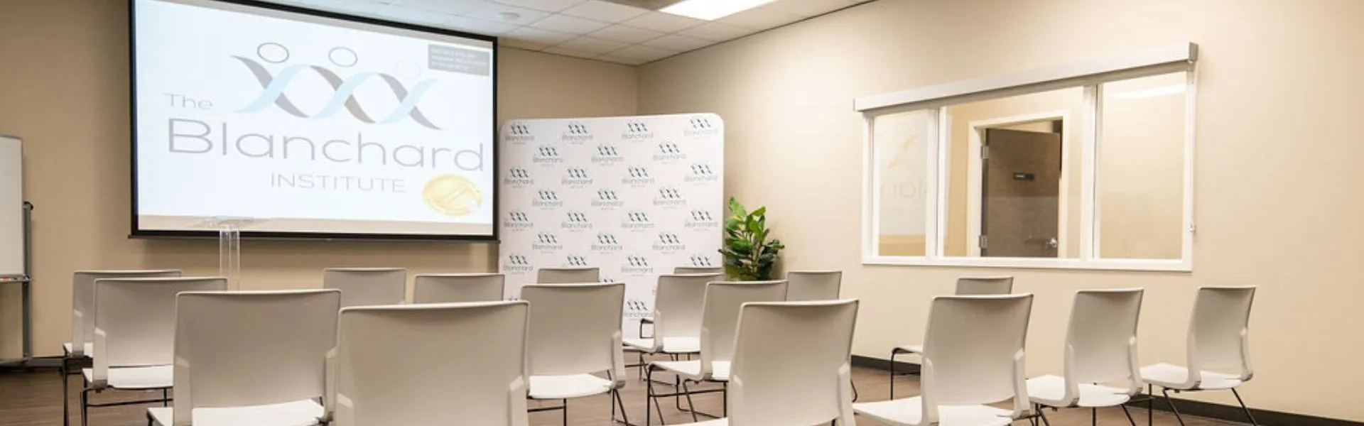 A small conference room at The Blanchard Institute, featuring rows of white chairs facing a projector screen. The screen displays the Blanchard logo. A step-and-repeat photo backdrop and a potted plant are placed in the corner, and there is a large window on the right wall.