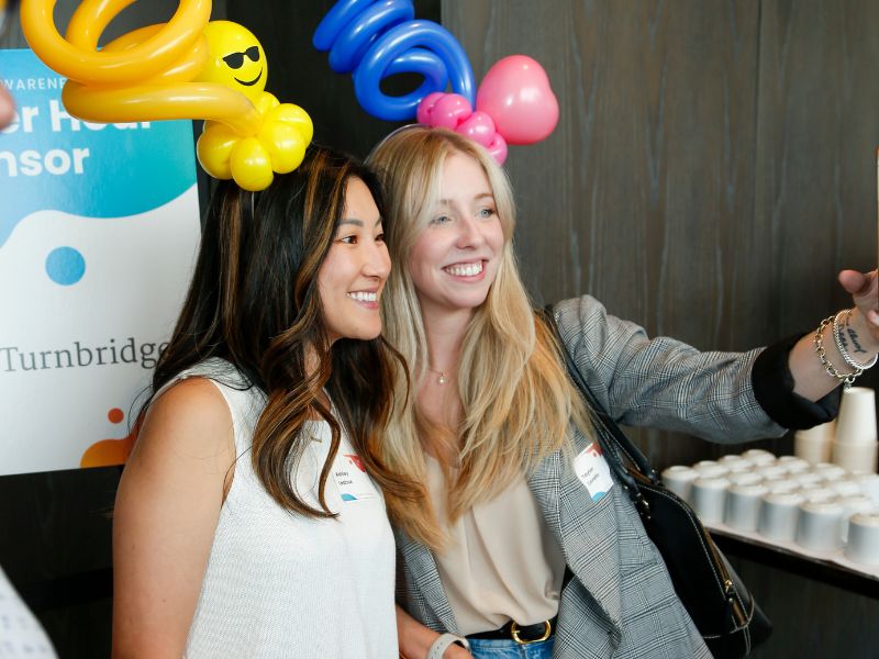 Two businesswomen taking a selfie at Recovery Awareness Day with colorful balloon hats and sponsor banners in the background.