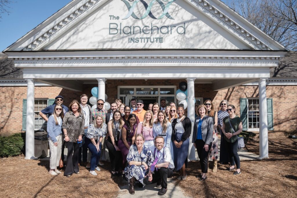 Large group photo of The Blanchard Institute team members and industry colleagues standing in front of the facility's main entrance with white columns, brick exterior, and balloons, celebrating the launch of the new Mental Health program.