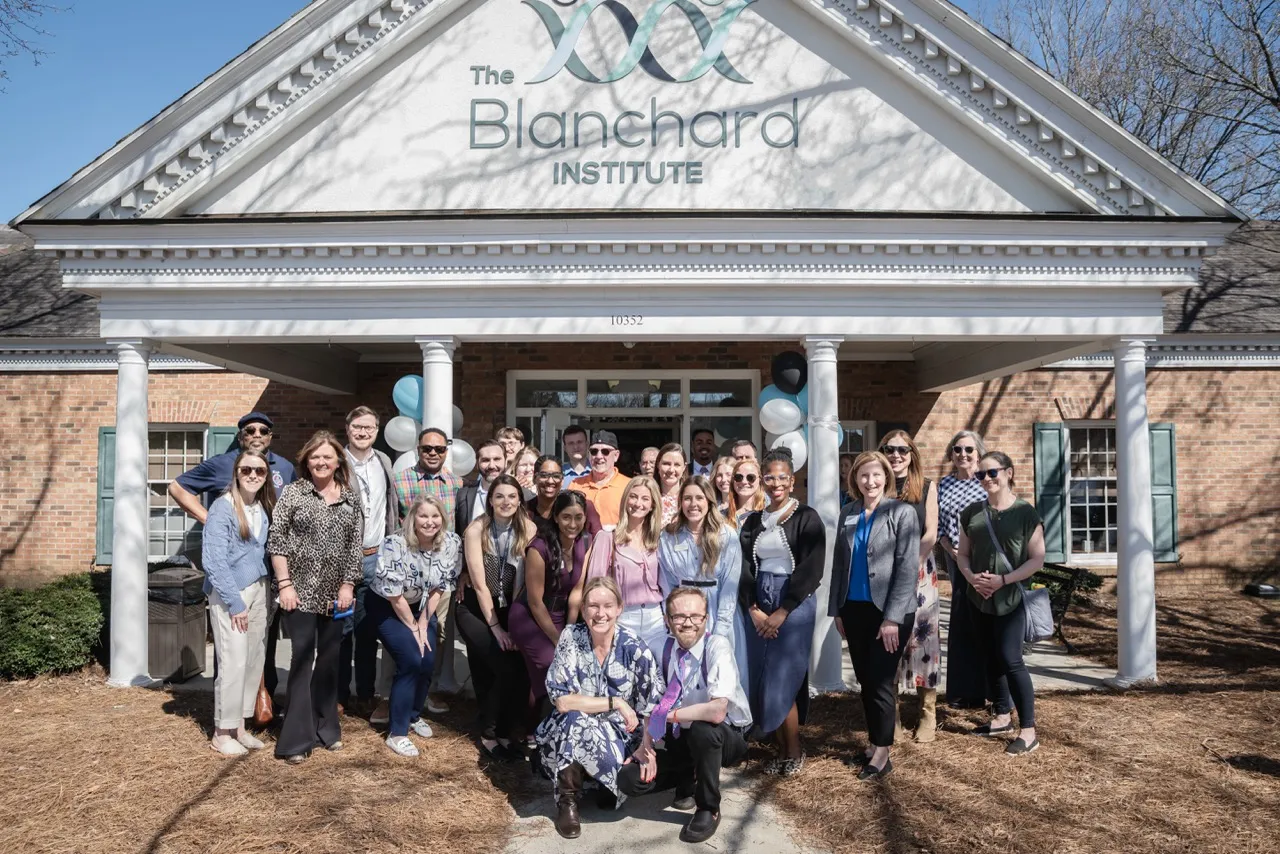 Large group photo of The Blanchard Institute team members and industry colleagues standing in front of the facility's main entrance with white columns, brick exterior, and balloons, celebrating the launch of the new Mental Health program.