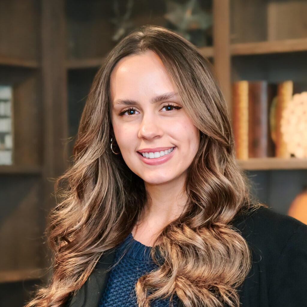 Professional headshot of a caucasian female with long wavy brown hair wearing business attire. She is standing in an office with decorated bookshelves behind her.