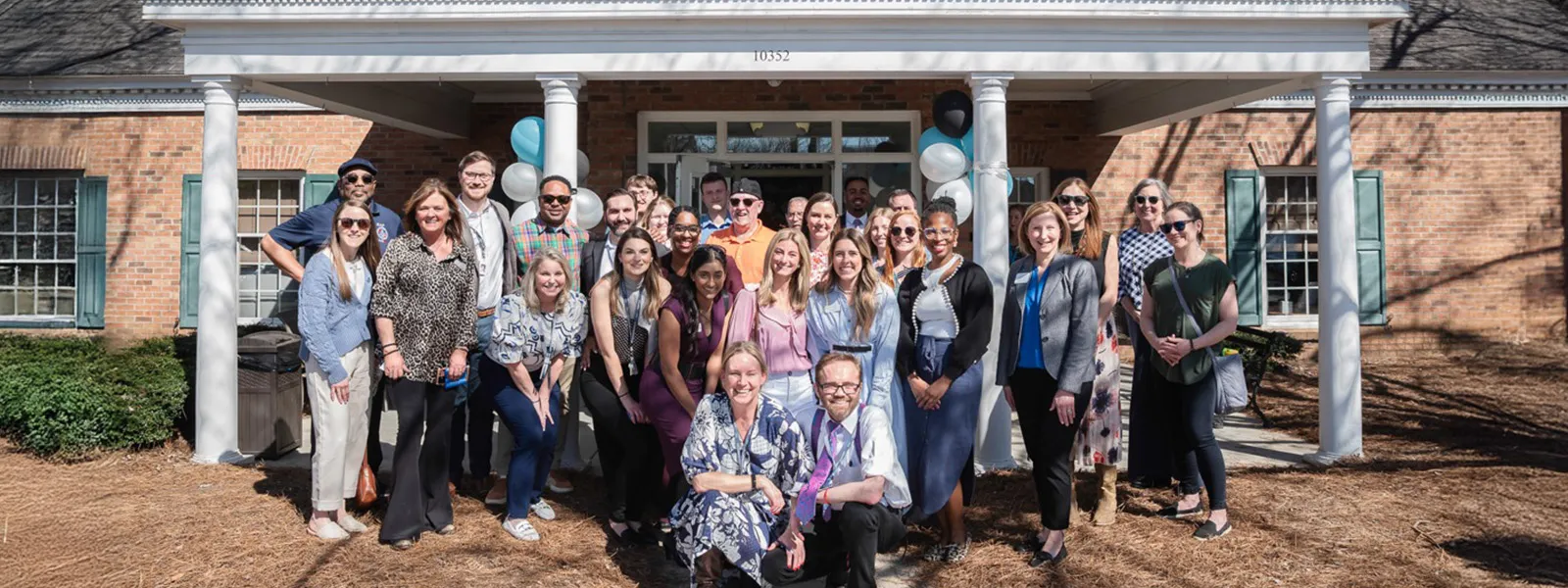 A large group of smiling staff members stands together outside The Blanchard Institute, posing in front of the main entrance on a sunny day. The team appears happy and connected, with balloons decorating the entryway, reflecting a warm and welcoming work culture.