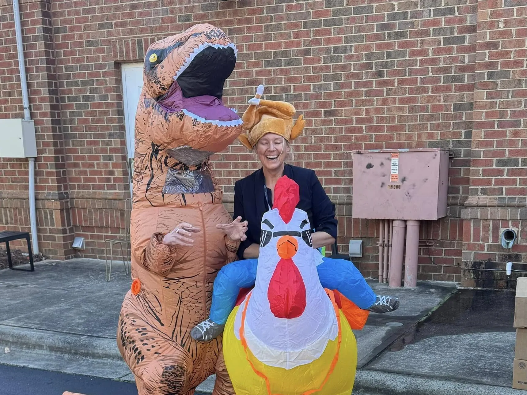 Two people in inflatable Halloween costumes posing playfully outside a brick building. One person wears a brown T-Rex dinosaur costume while the other sits in a colorful turkey costume with orange pilgrim hat.