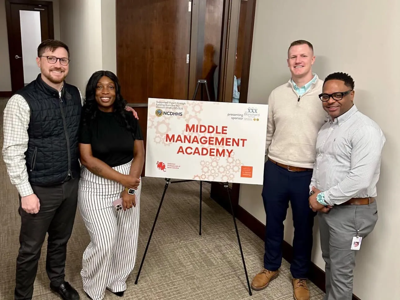 Four professional colleagues posing together around a presentation sign that reads 'Middle Management Academy' in an office conference room. The group includes two men in business casual attire on the left, a woman in professional striped clothing, and a man in a sweater vest on the right.